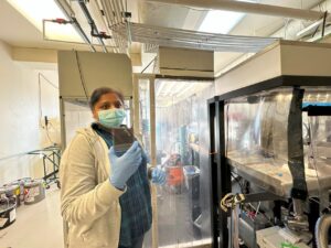Picture of a woman wearing a mask, gloves, and a lab coat, standing in a lab where several machines are draped in plastic sheeting