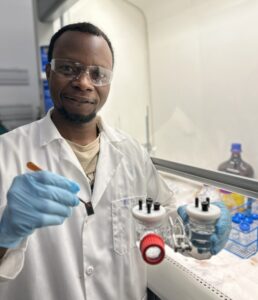 Picture of a student wearing goggles, gloves, and a lab coat, standing in front of a hood and holding tweezers and a small white device with black protrusions