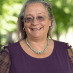 Picture of a woman smiling woman wearing a purple blouse, glasses, a green bead necklace, and small gold hoop earrings, standing in a location in nature